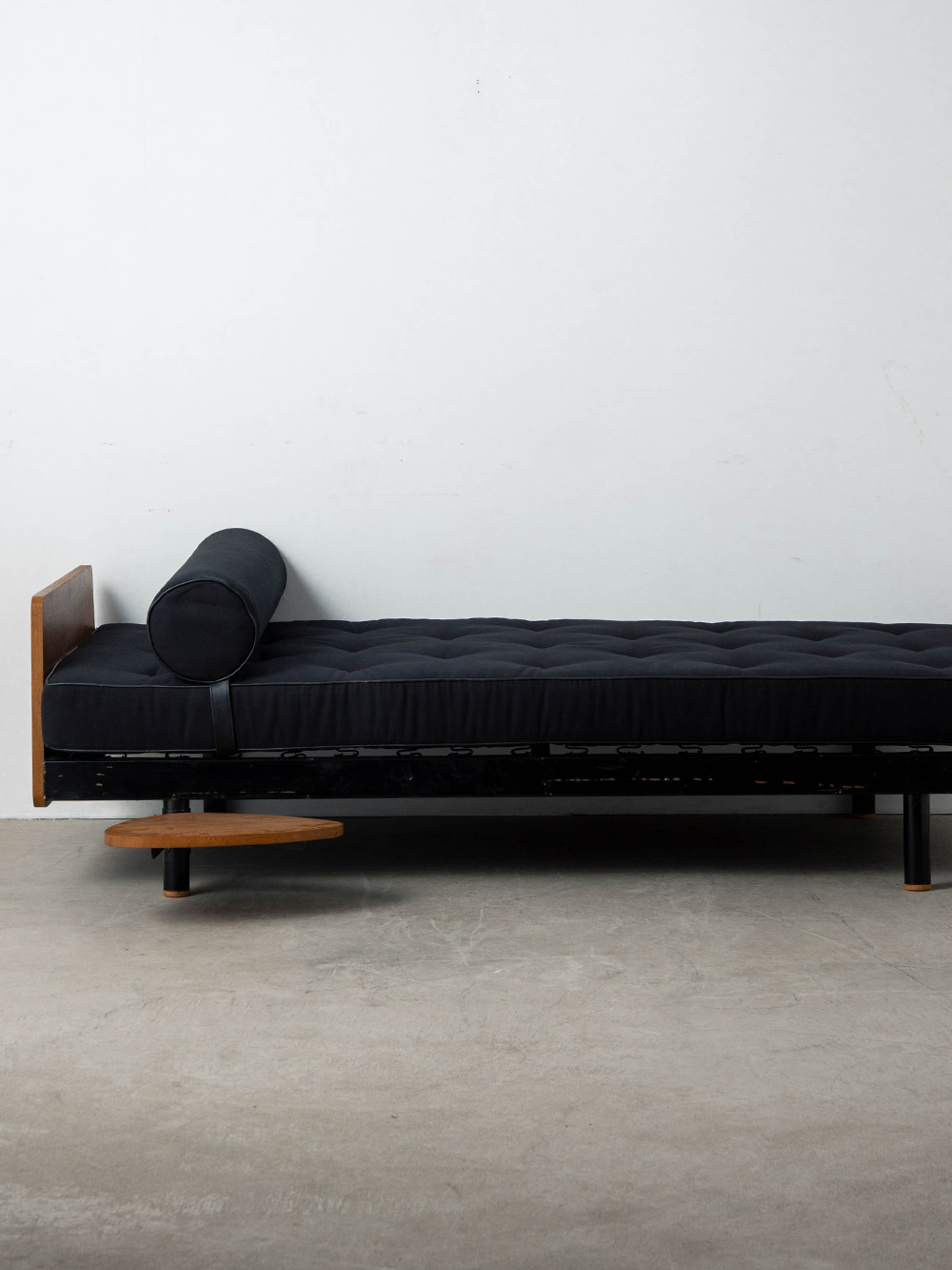 Scal Day Bed by Jean Prouvé , Charlotte Perriand in Steel and Wood
France , 1950s
フランスの建築家・デザイナーである Jean Prouvé（ジャン・プルーヴェ）と、同じくフランスを代表するデザイナー Charlotte Perriand（シャルロット・ペリアン）の協働によって生まれた SCAL デイベッド。
1940年に、名前の由来となる SCAL 工場従業員寮のために考案されたプルーヴェのモジュール式ベッドをベースに、1955年のパリ近郊アントニー大学都市の学生寮プロジェクトに向けて、プルーヴェとペリアンが協働しアップデートした希少なモデルです。
当時、学生寮の限られたスペースを最大限に活用するという課題に対し、室内装備の設計を任されていたペリアンは、プルーヴェの既存フレームに新たな機能を付加するアイデアを提案します。
それが、脚部に取り付けられた涙型の回転式サイドテーブルでした。
ベッドでありながら日中は壁付けにしてソファのように使用することができ、必要に応じて引き出せる木製テーブルが本やコーヒーカップを置くためのスペースとなります。
この合理的かつ温かみのあるディテールは、モジュール建築の構造原理を追求したプルーヴェの思想と、人々の暮らしに寄り添うペリアンの視点とが見事に融合したものです。
本個体は、アントニー大学都市プロジェクトに由来する通称「Antony Bed（アントニー・ベッド）」として知られるモデルで、パリの Steph Simon（ステフ・シモン）ギャラリーを通じて販売された希少な個体。
ヘッドボードとフットボードを備えた仕様であり、現存数の少ない貴重な一台です。
