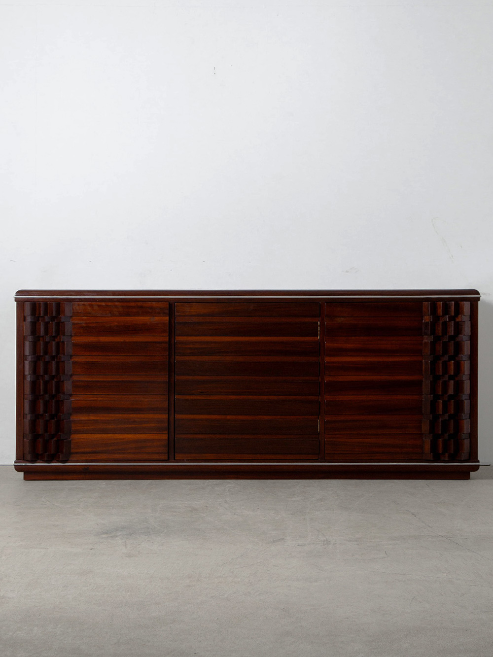 Diamante Sideboard by Luciano Frigerio for Frigerio di Desio in Wood
Italy , 1970s
イタリアのデザイナー Luciano Frigerio（ルチアーノ・フリジェリオ）によって1970年代にデザインされた Diamante（ディアマンテ）サイドボード。
両サイドにはジグザグ状の幾何学的なレリーフが施され、立体的な木部がリズミカルに連続することで、彫刻的な存在感を生み出しています。重厚感のある無垢材ならではの豊かな表情と深い色調が相まって、唯一無二の造形へと昇華しています。
1928年に家具工房を営む父のもとに生まれたフリジェリオは、その家業を発展させるかたちで Frigerio di Desio社を築き上げ、1960-70年代には、Mobili Scultura（家具彫刻）という独自の表現を追求しました。素材そのものの特性を重視した彼は、厳選された良質な無垢材を用いながら、工業的な均質さではなく、木の質感や重量感を積極的にデザインへ取り込み、独自の世界観を築き上げました。
